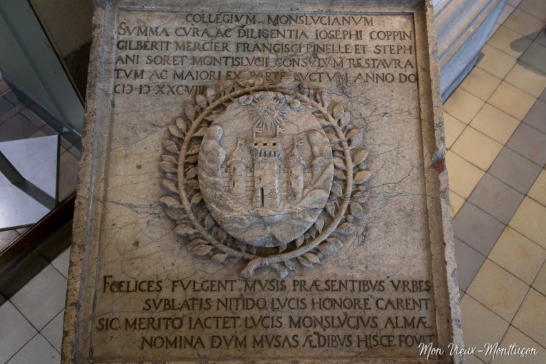 Hall de l'hôtel de ville : plaque de marbre qui était placée sur le fronton du nouveau collège de Montluçon en 1588.Traduction de l'inscription qui entoure les armoiries de la ville tirée du livre d'Henry Laguérenne Simple croquis de Montluçon au bon vieux temps (1904).Partie haute : Le collège de Montluçon, par les soins diligents et par le zèle de Joseph Coppin, Gilbert Mercier, François Pinel et Étienne Soret, consuls, a été restauré, et, en très grande partie, accru, cette année du Seigneur mil cinq cent quatre-vingt-huit.Partie basse : Heureuses les villes fréquentées par les Muses et parées de leur brillant éclat. Sans elles, cette lumière qui embellit et honore les cités leur est enlevée. C'est pourquoi Montluçon est à juste titre brillante de cette bienfaisante clarté, car, son nom l'indique, elle donne hospitalité aux Muses. 0113_mairie_marbre-armoirie.jpg